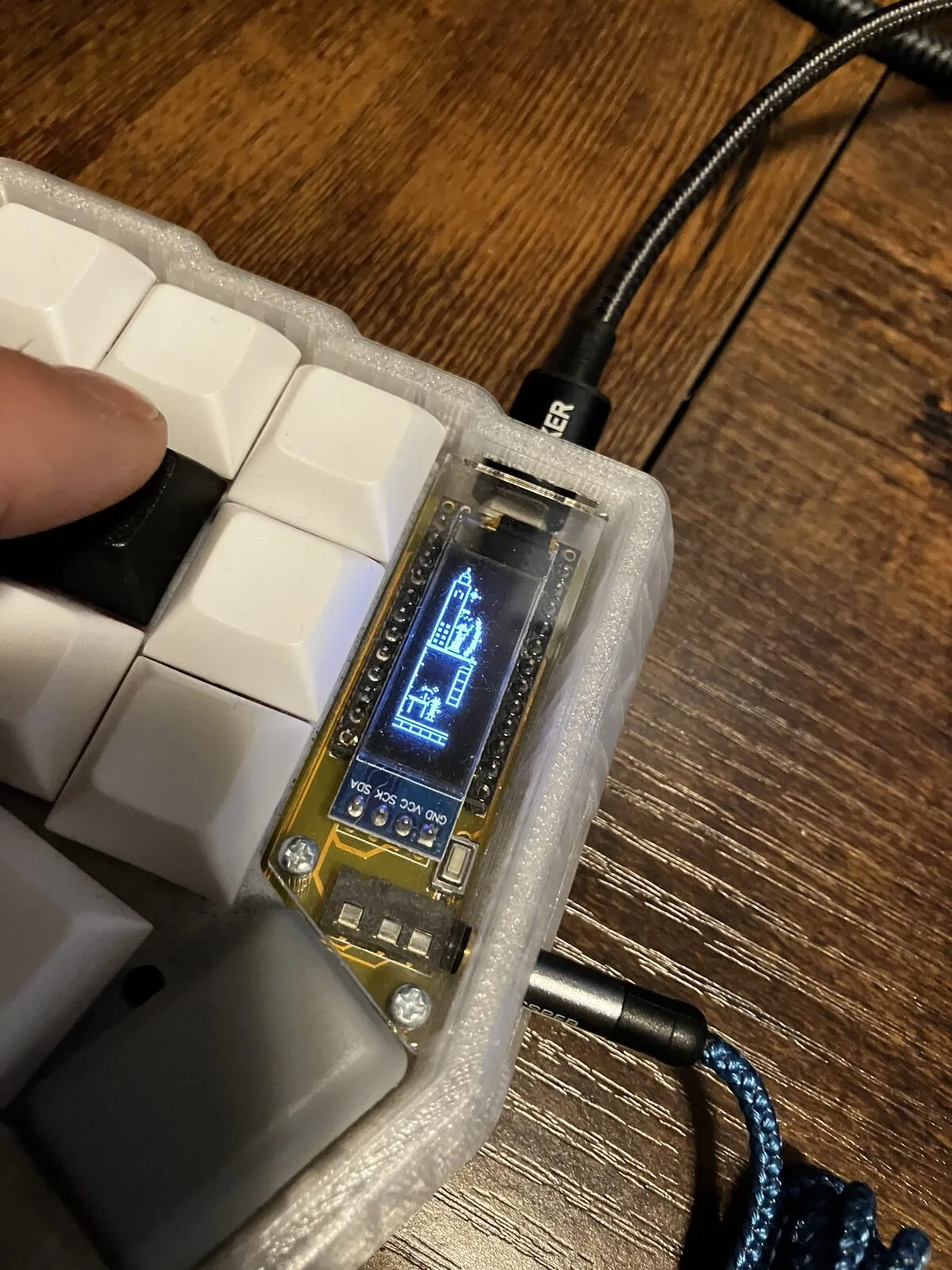 The master half of a Corne keyboard under USB and TRRS, its OLED lit blue and rendering a city skyline with tower silhouettes.